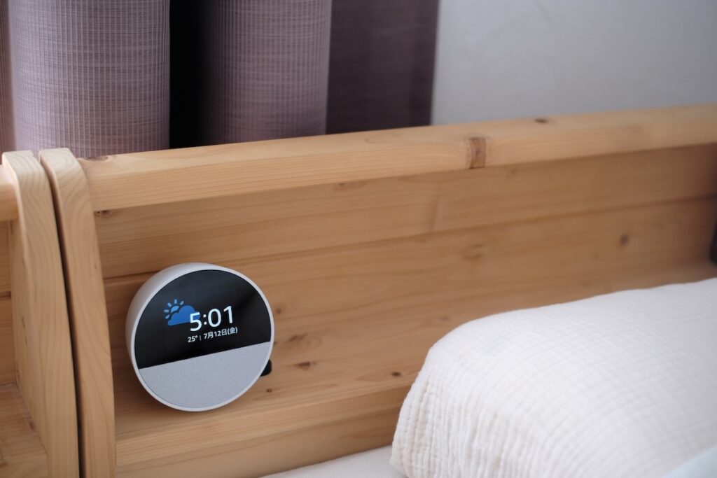ベッドのヘッドボードに設置されたEcho Spot(ホワイト)の実物写真(時刻5:01と天気アイコンが表示)
